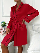 Femme portant un peignoir court en satin rouge vif, agrémenté de liserés noirs élégants, avec ceinture à nouer et col châle. Modèle raffiné et soyeux, parfait pour des moments de détente glamour ou une allure romantique à la maison.