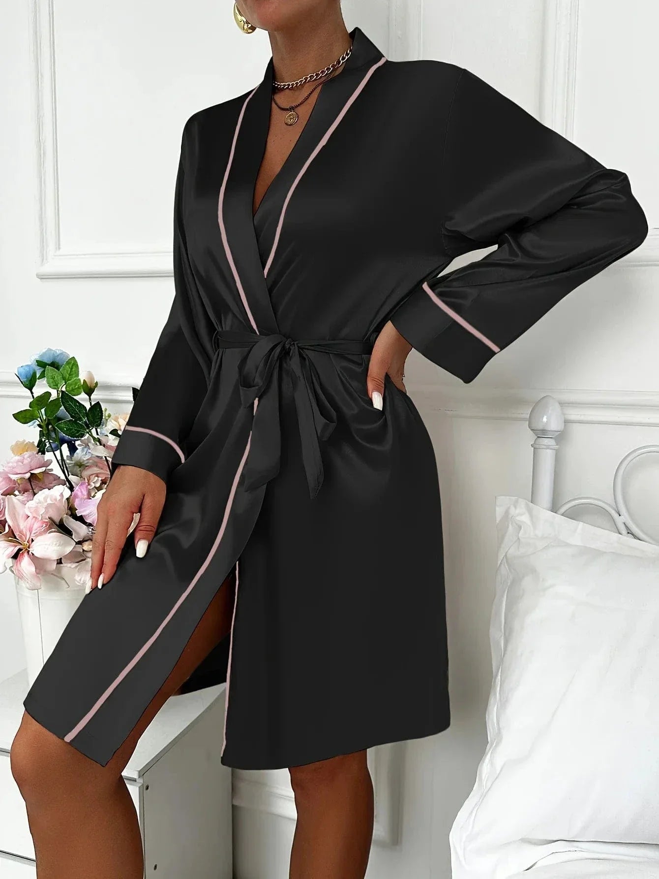 Femme portant un peignoir court en satin noir avec finitions rose pâle, col châle croisé et ceinture à nouer à la taille. Modèle élégant, doux au toucher, parfait pour les routines beauté, les matins raffinés ou comme idée cadeau féminine.