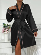Femme portant un peignoir court en satin noir orné de liserés rose pâle, avec ceinture à nouer et col châle. Modèle chic et confortable, parfait pour les instants de bien-être à la maison ou comme tenue élégante de nuit.