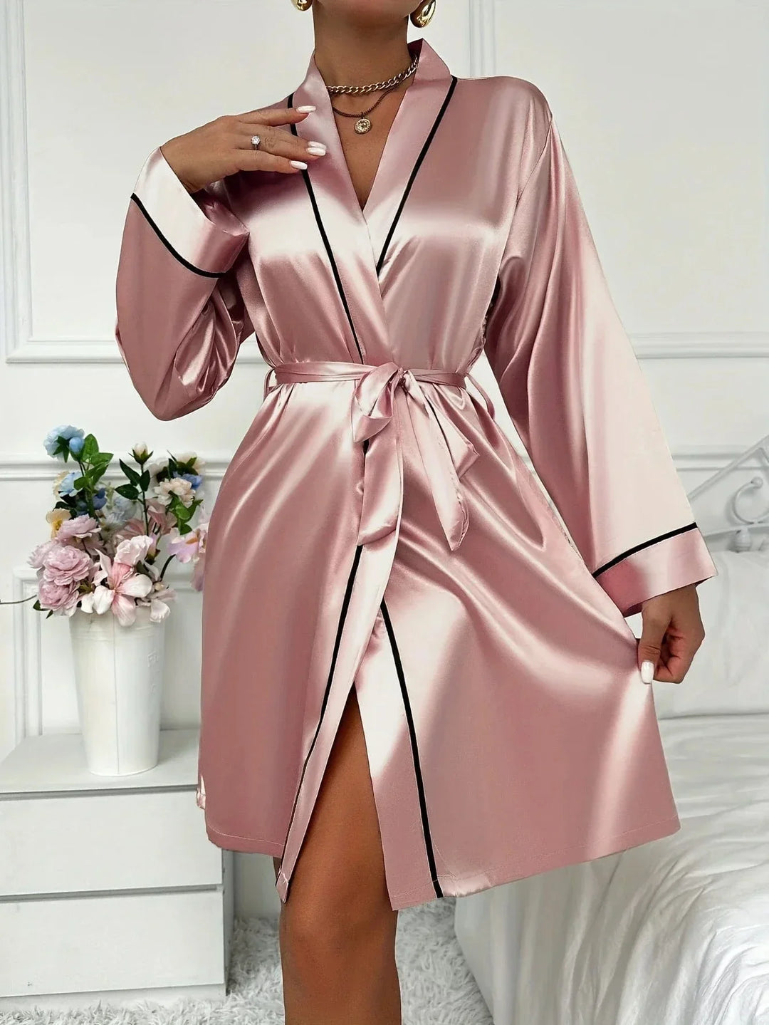 Femme portant un peignoir court en satin rose poudré avec des liserés noirs contrastants, col châle et ceinture à nouer. Modèle raffiné et soyeux, idéal pour une routine beauté élégante ou des moments de détente luxueux à la maison.