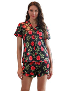 Femme portant le pyjama short femme, un ensemble léger et élégant idéal pour l’été, composé d’une chemise à manches courtes et d’un short assorti en satin noir imprimé de roses rouges et de feuilles vertes.