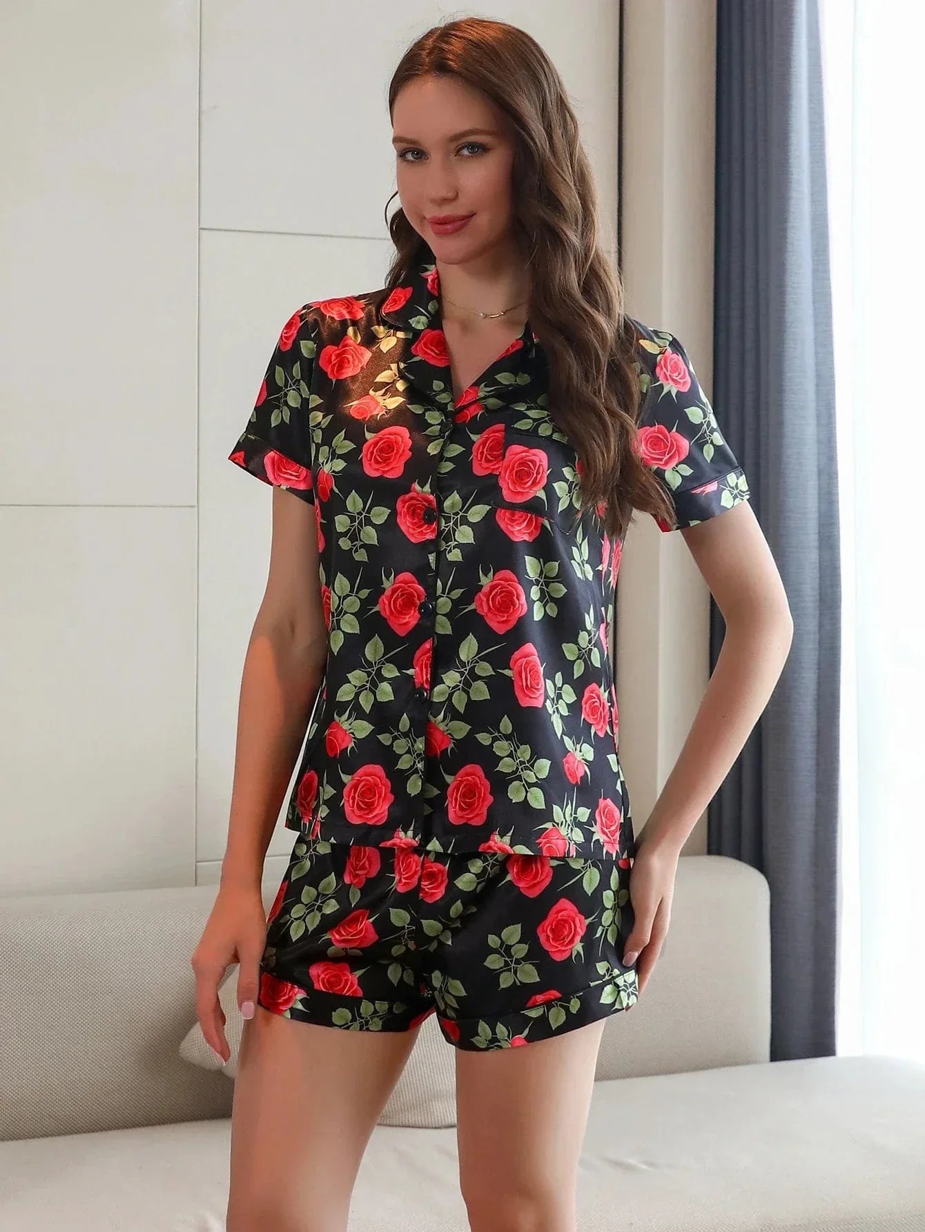 Femme portant le pyjama short femme, un ensemble léger et élégant parfait pour l’été, composé d’une chemise à manches courtes et d’un short assorti en satin noir orné d’un imprimé de roses rouges et de feuilles vertes.