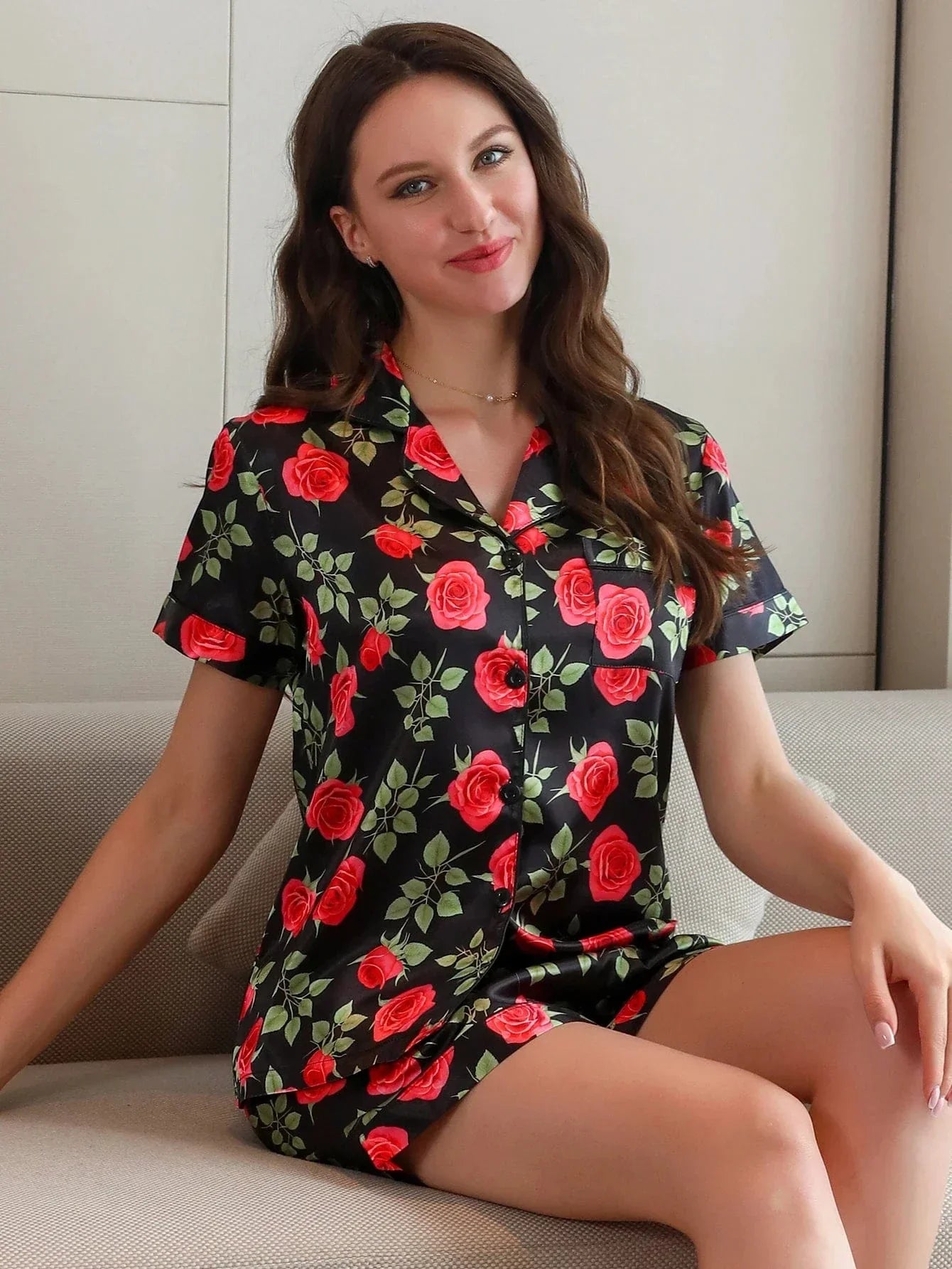 Femme assise portant le pyjama short femme, un ensemble léger et élégant parfait pour l’été, composé d’une chemise à manches courtes et d’un short assorti en satin noir imprimé de roses rouges et de feuilles vertes.