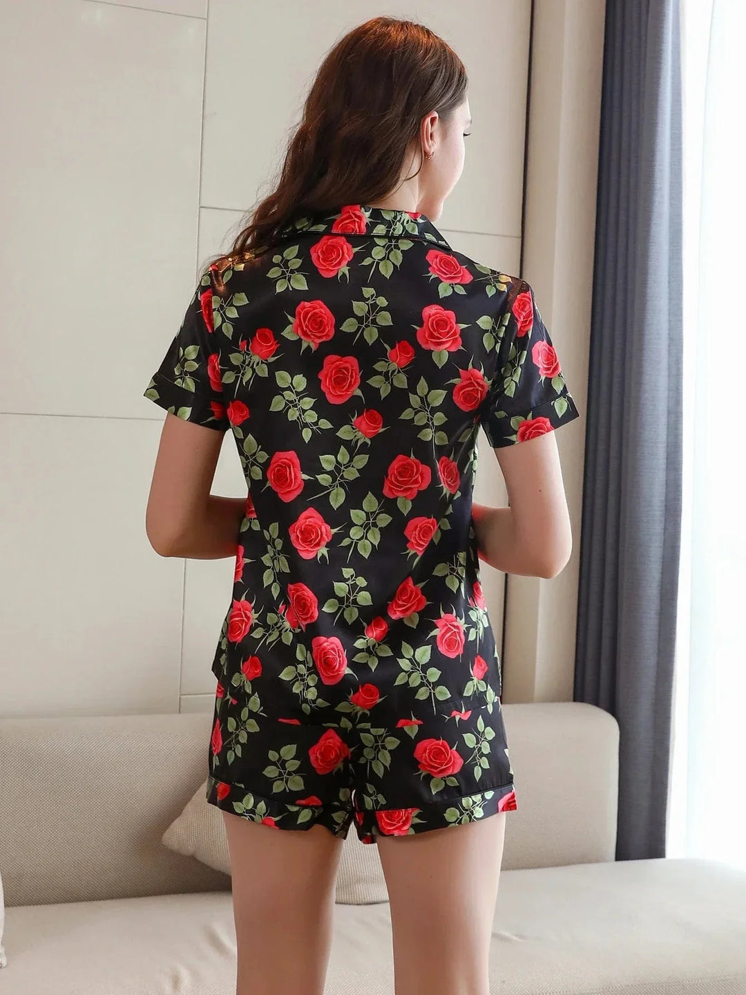 Femme vue de dos portant le pyjama short femme, un ensemble léger et élégant parfait pour l’été, composé d’une chemise à manches courtes et d’un short assorti en satin noir décoré d’un imprimé de roses rouges et de feuilles vertes.