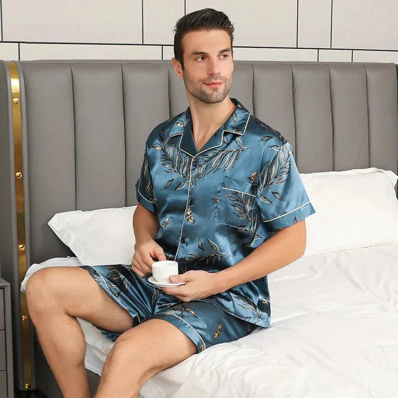Homme assis sur un lit portant le shorty satin François pour homme, ensemble deux pièces avec chemise boutonnée à manches courtes et short assorti, motif feuillage sur fond bleu-vert, vue de face, présenté en intérieur.