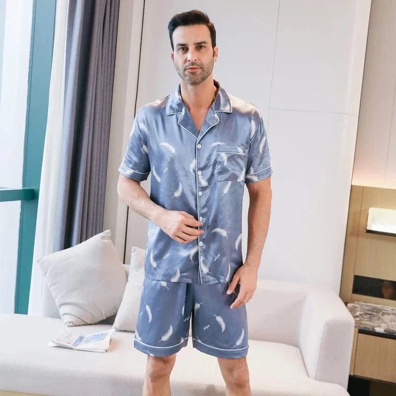 Homme debout portant le shorty satin Gabriel pour homme, ensemble deux pièces avec chemise boutonnée à manches courtes et short assorti, motif plume clair sur fond bleu, coupe droite, présenté en intérieur.