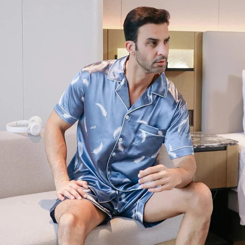 Homme assis portant le shorty satin Gabriel pour homme, ensemble deux pièces avec chemise boutonnée à manches courtes et short assorti, motif plume clair sur fond bleu, vue de trois-quarts, présenté en intérieur.