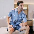 Homme assis portant le shorty satin Gabriel pour homme, ensemble deux pièces avec chemise boutonnée à manches courtes et short assorti, motif plume clair sur fond bleu, vue de trois-quarts, présenté en intérieur.