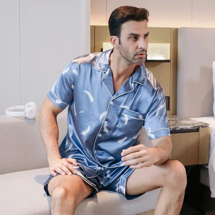 Homme assis portant le shorty satin Gabriel pour homme, ensemble deux pièces avec chemise boutonnée à manches courtes et short assorti, motif plume clair sur fond bleu, vue de trois-quarts, présenté en intérieur.