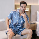 Homme assis portant le shorty satin Gabriel pour homme, ensemble deux pièces avec chemise boutonnée à manches courtes et short assorti, motif plume clair sur fond bleu, vue de face, présenté en intérieur.