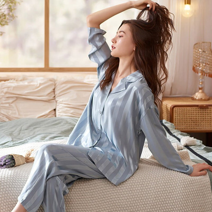 Modèle assise sur un lit portant le pyjama femme Garance bleu satiné à rayures, chemise boutonnée manches longues et pantalon assorti, tissu doux et confortable dans une chambre lumineuse.