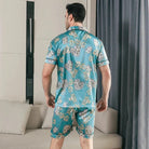 Vue arrière d’un homme portant le pyjama satin Georges pour homme, ensemble deux pièces avec chemise à manches courtes et short assorti, imprimé décoratif vert turquoise, coupe droite, présenté en intérieur.