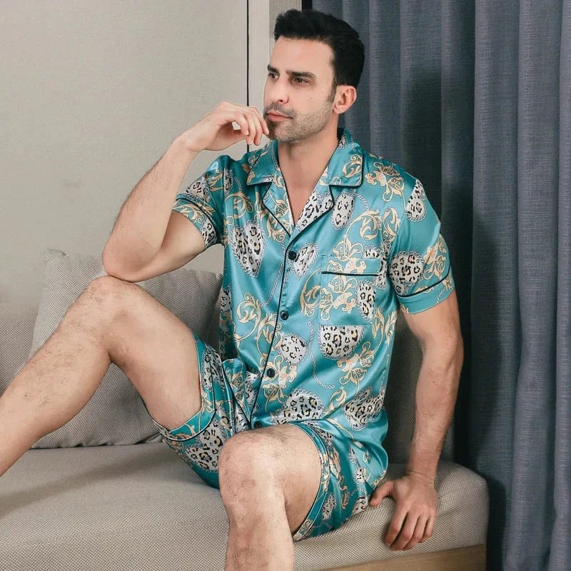 Homme assis portant le pyjama satin Georges pour homme, ensemble deux pièces avec chemise boutonnée à manches courtes et short assorti, imprimé décoratif vert turquoise, vue de trois-quarts, présenté en intérieur.