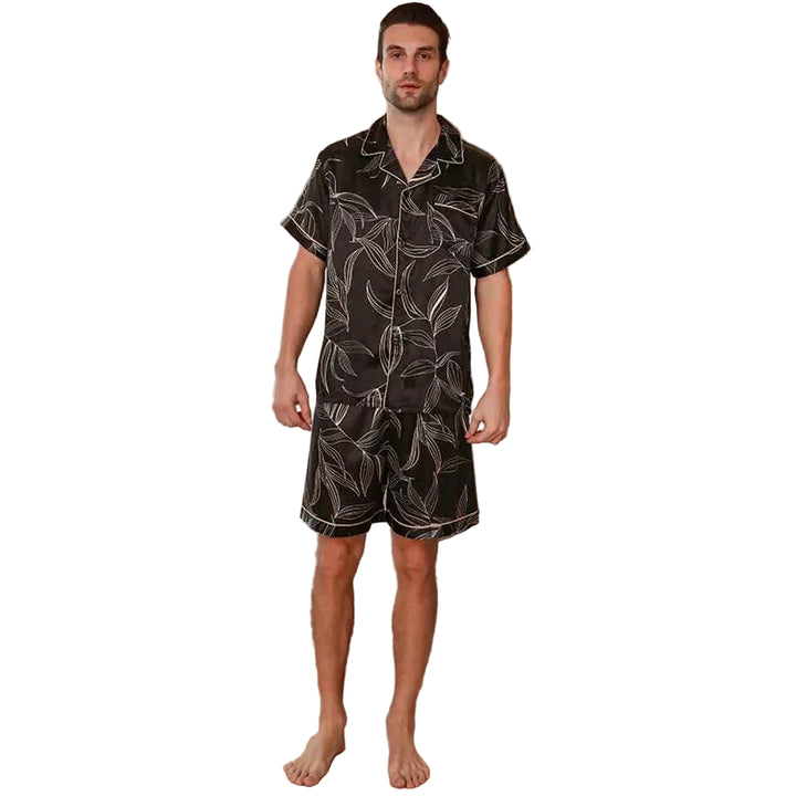 Homme debout pieds nus dans un intérieur moderne portant le shorty satin pour homme Guillaume, ensemble élégant et agréable composé d’une chemise à manches courtes et d’un short assorti, en satin noir à imprimé feuilles, offrant une allure raffinée et un confort idéal pour la maison ou la nuit.