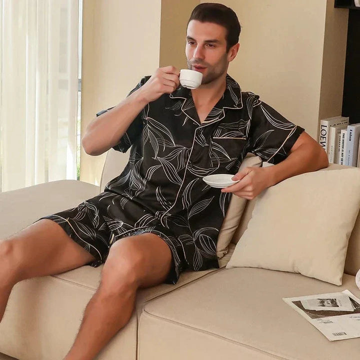 Homme assis sur un canapé tenant une tasse, portant le shorty satin pour homme Guillaume, ensemble élégant et agréable composé d’une chemise à manches courtes et d’un short assorti, en satin noir à imprimé feuilles, offrant un style raffiné et un confort idéal pour la détente à la maison.