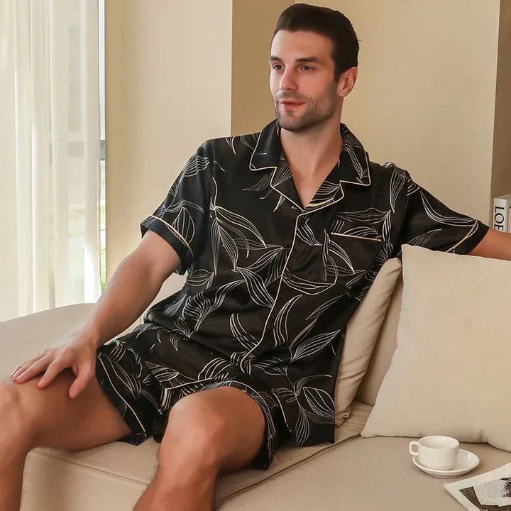 Homme assis sur un canapé portant le shorty satin pour homme Guillaume, ensemble élégant et agréable en satin noir à imprimé feuilles, composé d’une chemise à manches courtes et d’un short assorti, offrant une allure raffinée et un confort optimal pour la détente à la maison.