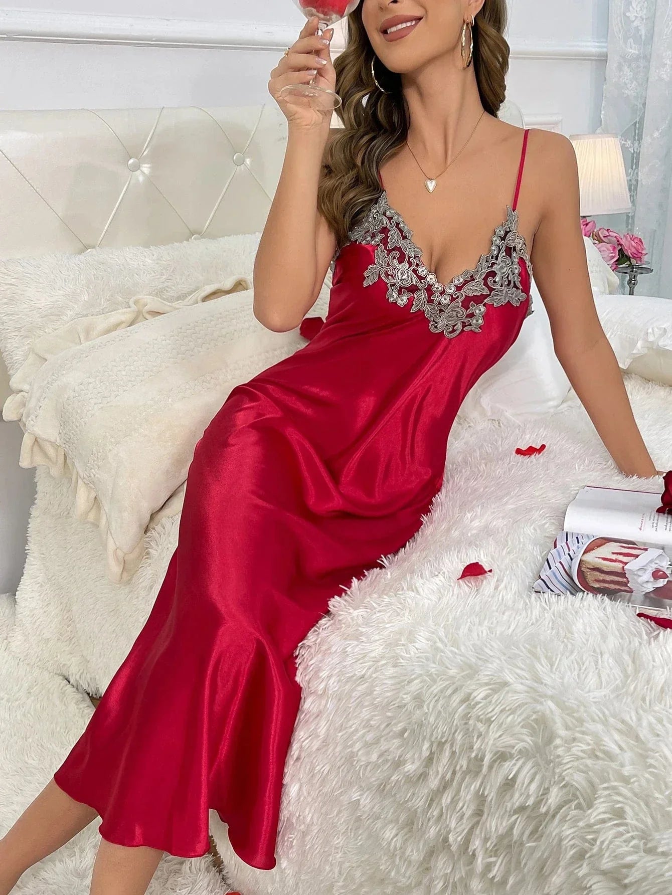 Femme assise sur un lit, souriante, portant le peignoir long Héloïse en satin rouge avec détails en dentelle grise aux poignets, tenant un verre à vin. Lingerie élégante et agréable à porter, parfaite pour les instants de détente ou les occasions spéciales à la maison.