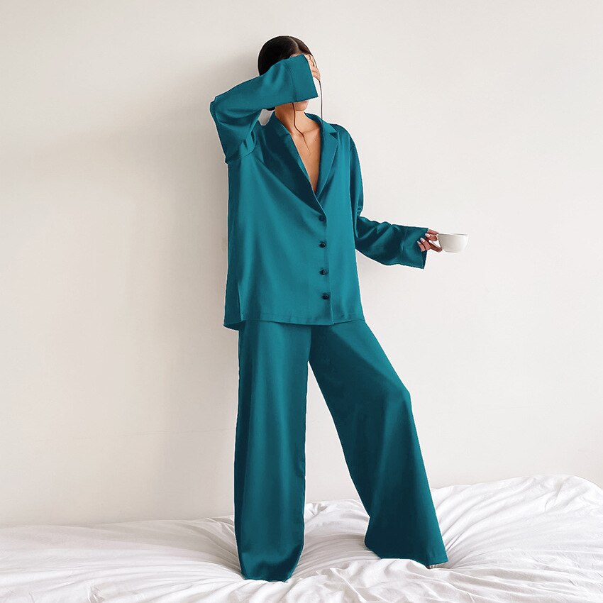 Femme debout sur un lit, portant un pyjama satiné vert émeraude composé d’une chemise fluide boutonnée à manches longues et d’un pantalon ample. Ensemble léger et élégant, idéal pour un style décontracté et sophistiqué à la maison.