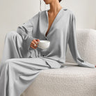 Femme assise sur un fauteuil texturé, vêtue d’un pyjama satiné gris clair composé d’une chemise fluide à boutons et manches longues, et d’un pantalon ample. Ensemble chic, léger et confortable, parfait pour un look raffiné à la maison.