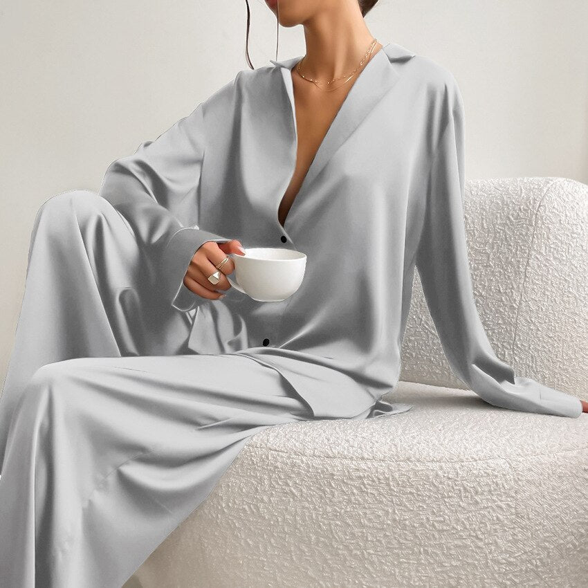 Femme assise sur un fauteuil texturé, vêtue d’un pyjama satiné gris clair composé d’une chemise fluide à boutons et manches longues, et d’un pantalon ample. Ensemble chic, léger et confortable, parfait pour un look raffiné à la maison.