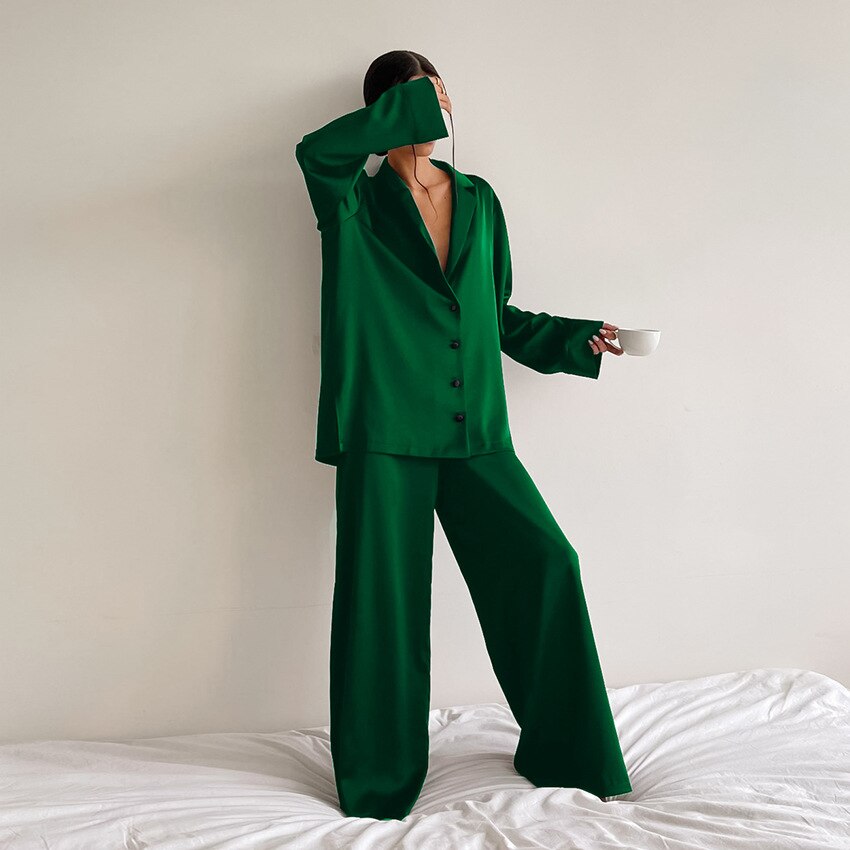 Femme debout sur un lit, vêtue d’un pyjama satiné vert foncé composé d’une chemise boutonnée fluide à manches longues et d’un pantalon ample. Ensemble confortable et élégant, parfait pour un look chic à la maison ou en mode détente.