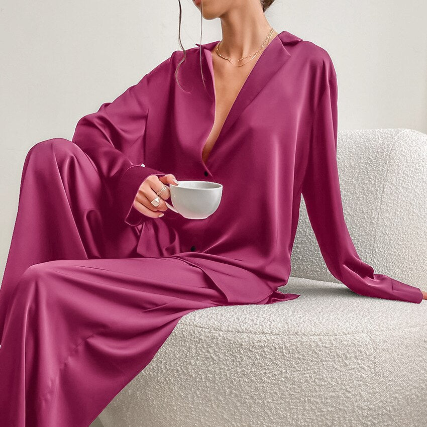 Femme assise dans un fauteuil, tenant une tasse, vêtue d’un pyjama satiné rose framboise composé d’une chemise fluide boutonnée et d’un pantalon ample. Ensemble léger, élégant et confortable, parfait pour des moments de détente avec style.
