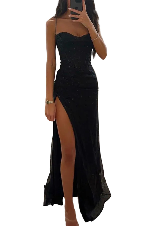 Une femme portant une robe maxi noire en satin avec une fente élégante sur le côté. La robe, décorée de détails scintillants, est idéale pour les occasions spéciales, offrant une silhouette sophistiquée et chic. Elle porte des accessoires raffinés et pose dans une chambre moderne, créant un look à la fois élégant et glamour.