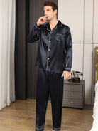 Pyjama en satin pour homme de couleur noire, avec une chemise à manches longues et une poche avant, assortie d’un pantalon. Le pyjama présente des finitions élégantes avec des bordures blanches sur le col, les poignets et le bas du pantalon. Le modèle est debout, en train de réfléchir, dans un cadre moderne et épuré. Ce pyjama est parfait pour allier confort et style lors de vos moments de détente à la maison.