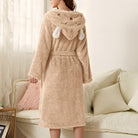 Peignoir femme chaud Iris en tissu peluche beige, coupe mi-longue avec ceinture, capuche fantaisie avec petites oreilles, porté de dos dans un salon.
