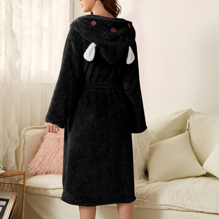 Peignoir femme chaud Iris en tissu peluche noir, coupe mi-longue avec ceinture, capuche fantaisie avec petites oreilles, porté de dos dans un salon.
