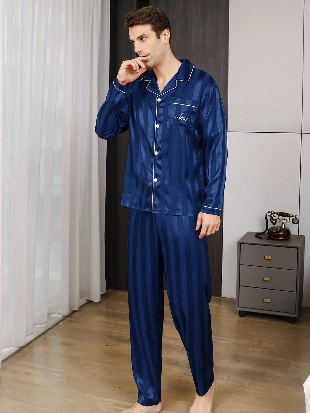 Homme debout dans un intérieur élégant portant le pyjama satin pour homme Jérôme, ensemble élégant et confortable composé d’une chemise à manches longues boutonnée et d’un pantalon assorti, en satin bleu à fines rayures avec liserés contrastés, offrant une allure raffinée et un confort optimal pour la nuit ou la détente à la maison.