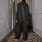 Une femme vue de dos dans un couloir élégant, portant un pyjama satiné noir à rayures verticales. Le contraste des rayures brillantes donne au tissu une allure sophistiquée.