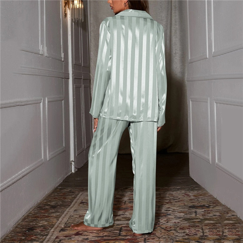 Une femme vue de dos dans un couloir élégant, portant un pyjama satiné vert tendre à rayures verticales. Le satin lumineux met en valeur la texture douce et soyeuse du vêtement.