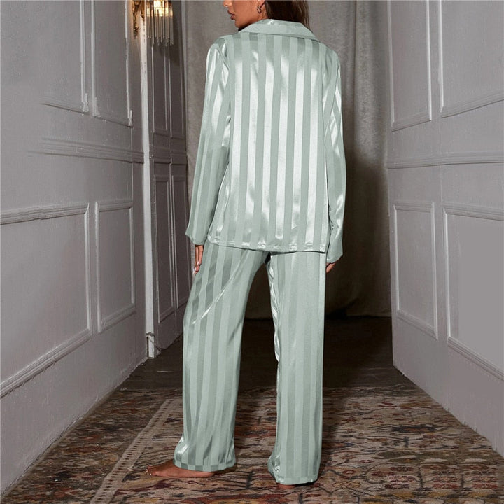 Une femme vue de dos dans un couloir élégant, portant un pyjama satiné vert tendre à rayures verticales. Le satin lumineux met en valeur la texture douce et soyeuse du vêtement.
