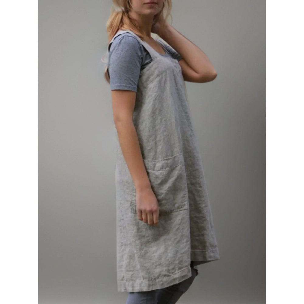 Vue de profil du tablier robe femme  pratique et confortable, en lin gris clair, coupe longue et droite avec bretelles larges, porté sur un t-shirt et un legging gris.