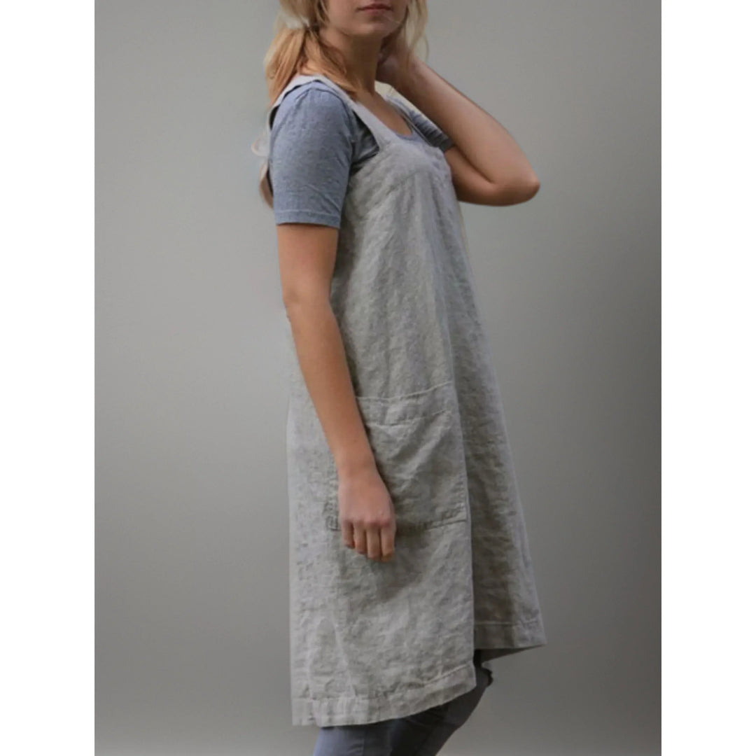 Vue de profil du tablier robe femme  pratique et confortable, en lin gris clair, coupe longue et droite avec bretelles larges, porté sur un t-shirt et un legging gris.