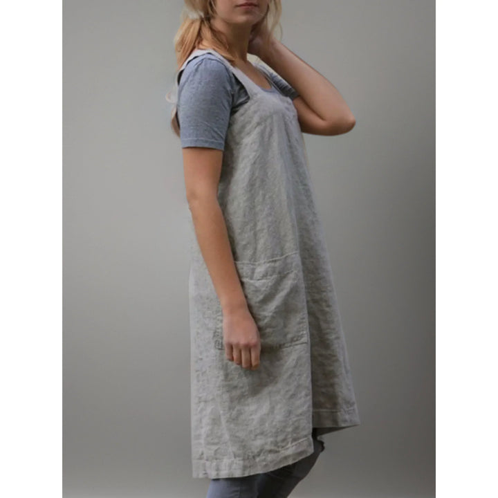 Vue de profil du tablier robe femme  pratique et confortable, en lin gris clair, coupe longue et droite avec bretelles larges, porté sur un t-shirt et un legging gris.