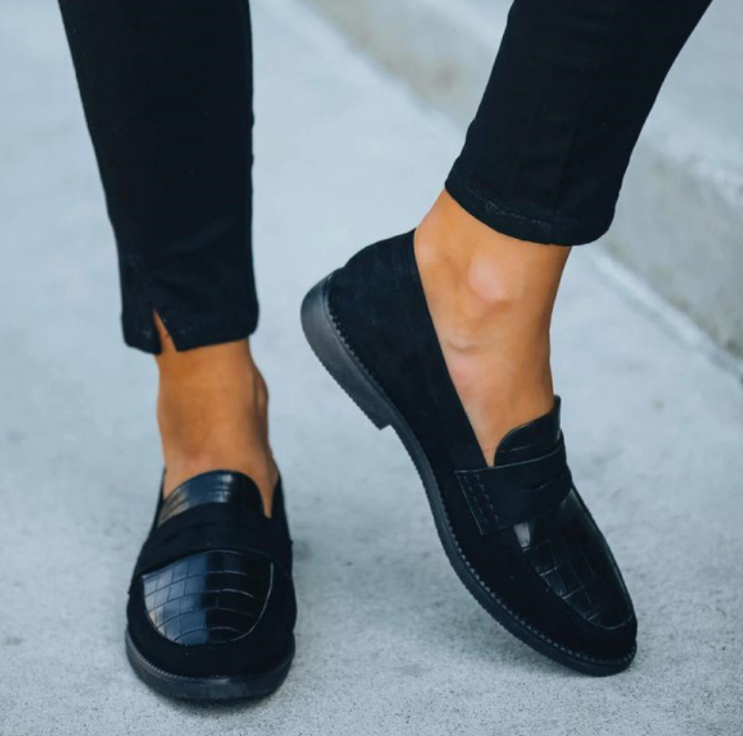 Mocassins femme élégants et confortables noirs, empiècement effet croco et bandeau, portés avec pantalon noir, vue de face avec un pied levé.
