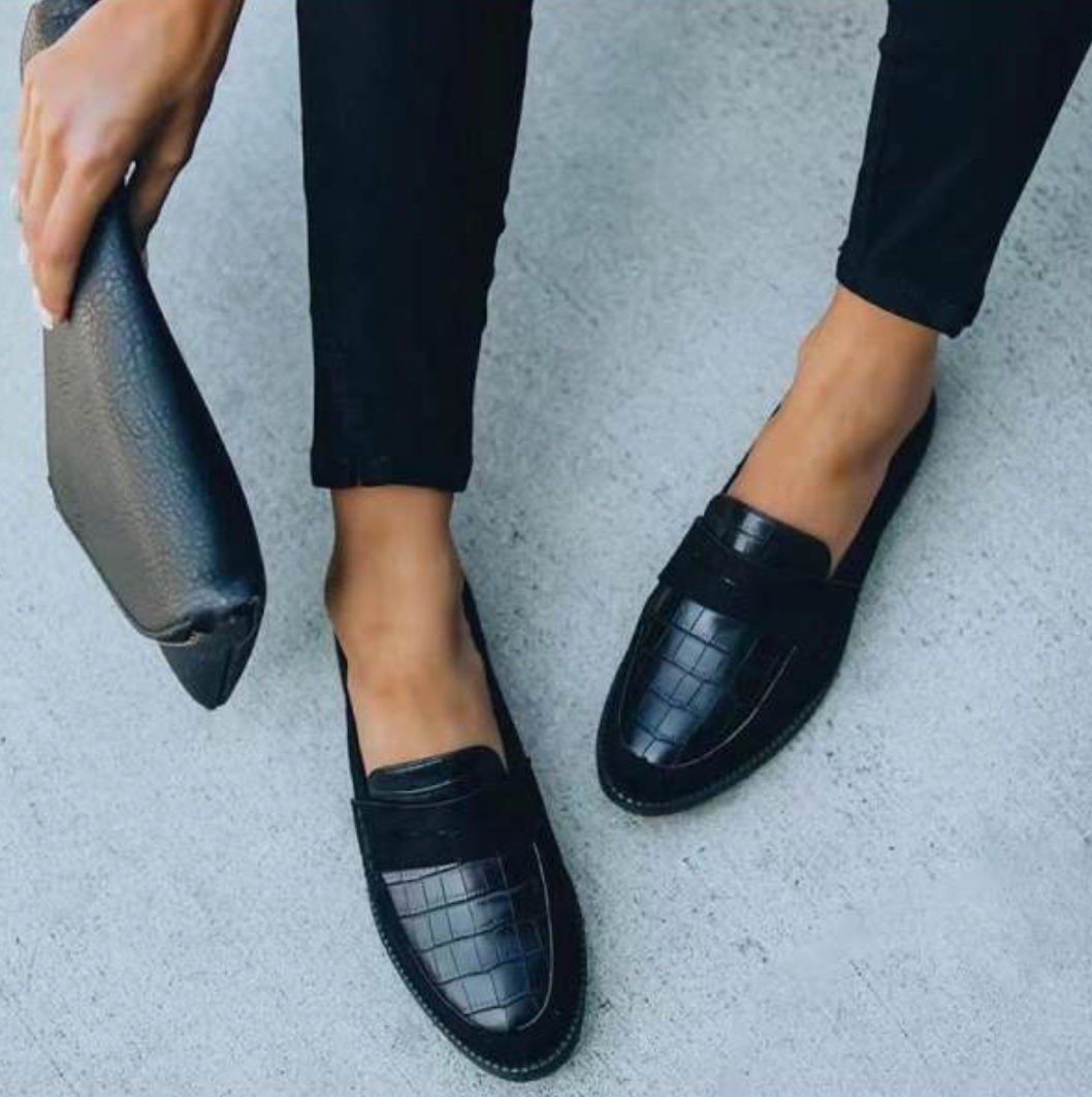 Mocassins femme élégants et confortables noirs, dessus effet croco avec bandeau, portés avec pantalon noir, photo lifestyle avec pochette à la main.