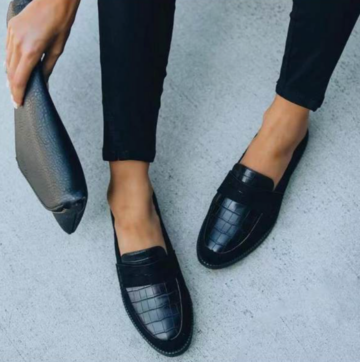 Mocassins femme élégants et confortables noirs, dessus effet croco avec bandeau, portés avec pantalon noir, photo lifestyle avec pochette à la main.