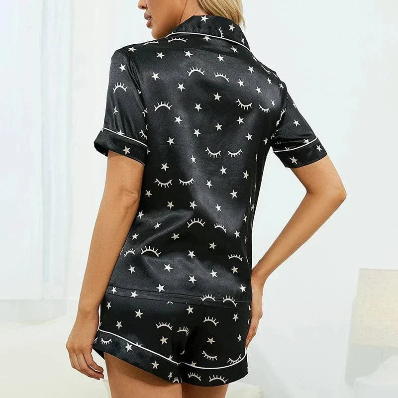 Vue de dos d'une femme portant le pyjama en satin pour femme. Léger, élégant et parfait pour la nuit, cet ensemble en satin noir se compose d'une chemise à manches courtes et d'un short, orné d'étoiles et de motifs de cils, offrant confort et style pour la nuit.