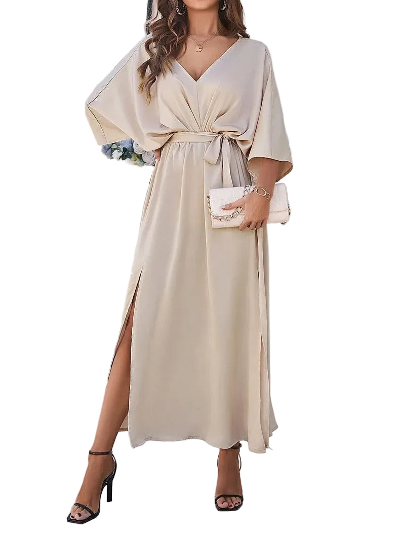 Femme portant la robe midi 'Katia', de couleur beige, avec un décolleté en V et une taille cintrée par un lien. Le tissu fluide et léger crée une silhouette élégante, avec des fentes sur les côtés pour une touche de modernité. Idéale pour les journées printanières, elle est complétée par une pochette et des sandales à talons.