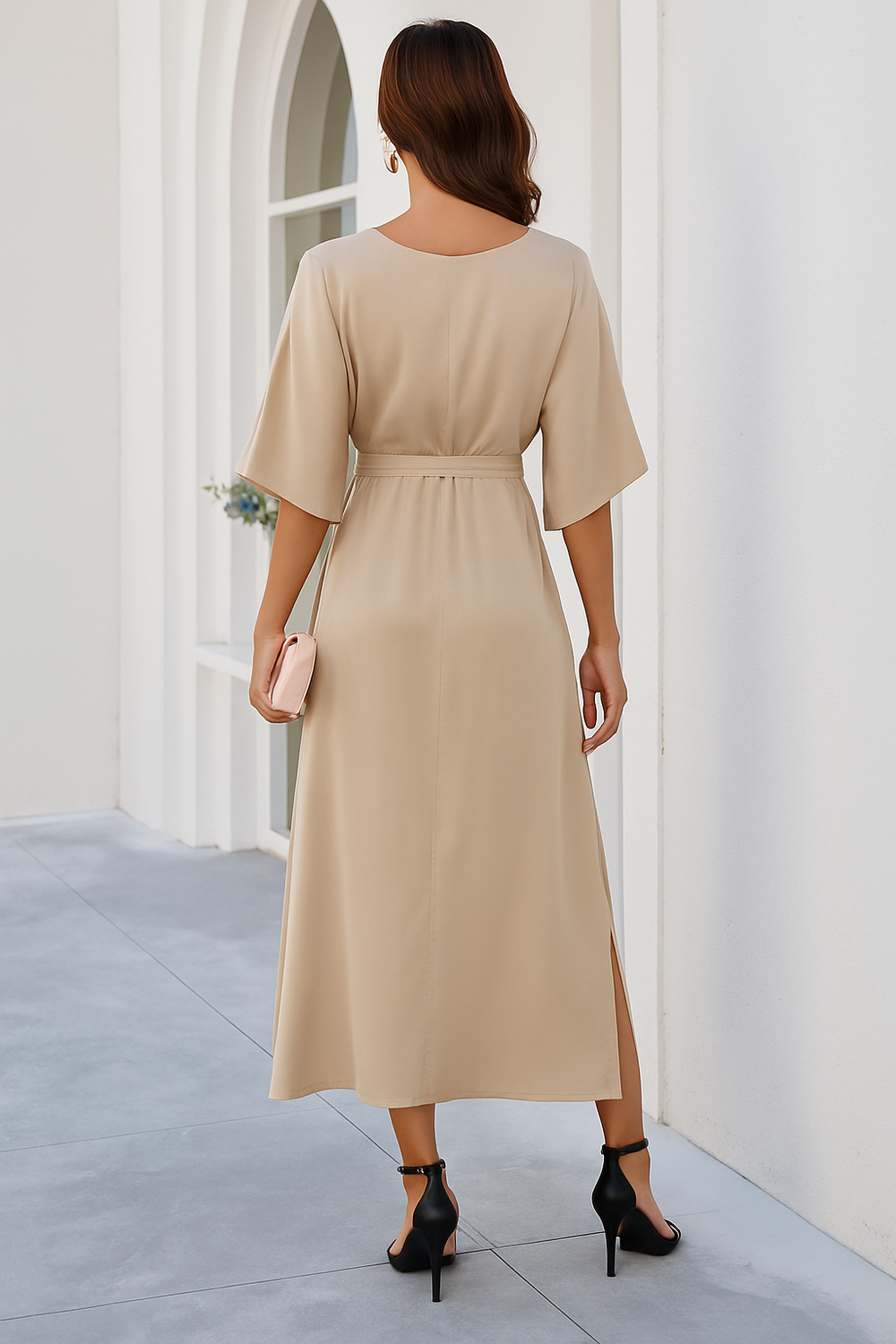 Vue arrière de la robe midi 'Katia', de couleur beige, avec un dos épuré et un lien à la taille pour marquer la silhouette. La robe à manches kimono et fentes latérales offre une allure élégante et fluide, idéale pour le printemps. La femme porte des sandales à talons et une pochette, parfaite pour une occasion spéciale ou une sortie élégante.