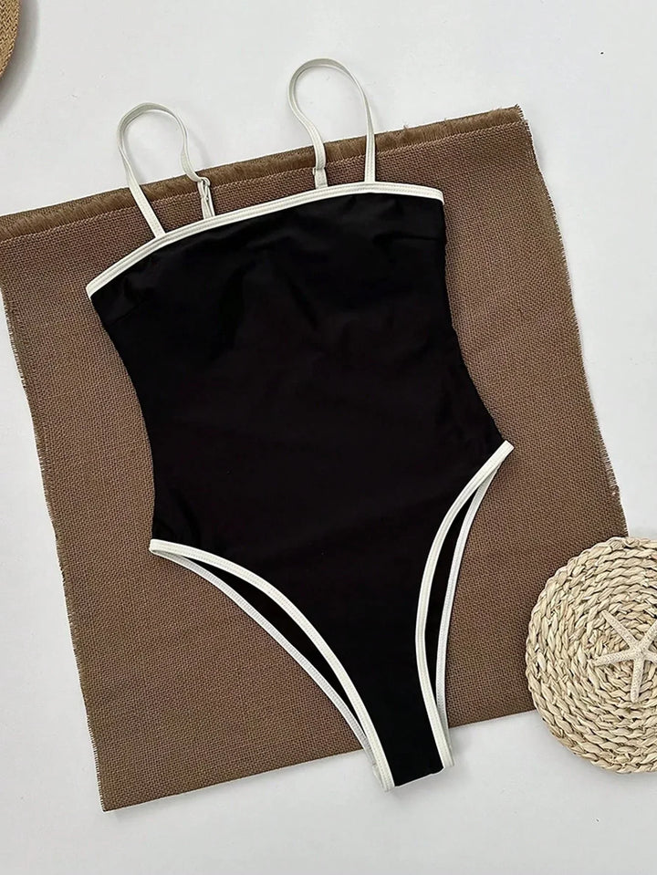 Maillot de bain femme une pièce noir avec bordures blanches contrastées et fines bretelles, présenté à plat sur un tissu texturé pour un style élégant, moderne et confortable.