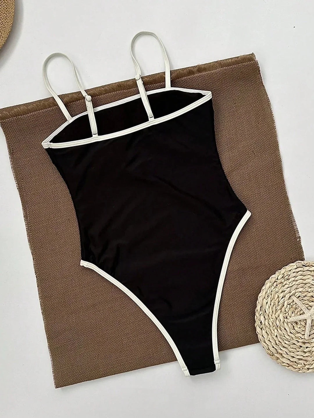 Maillot de bain femme une pièce noir avec bordures blanches contrastées et fines bretelles, présenté à plat sur un tissu texturé, vue arrière pour un style élégant, moderne et confortable.