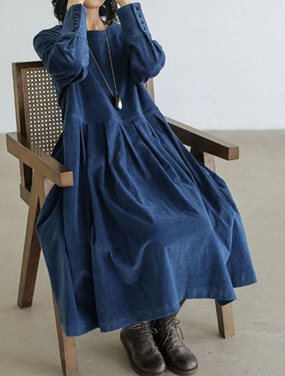 Robe femme élégante et raffinée, bleu marine, coupe ample et fluide avec manches longues boutonnées, portée assise sur une chaise en bois avec des bottines.
