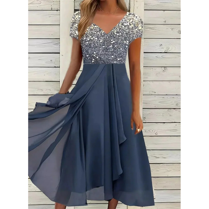 Robe femme élégante et polyvalente, longue et fluide, jupe bleu marine avec haut scintillant argenté, col en V et manches courtes, portée devant un mur en bois blanc.