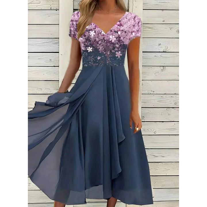 Robe femme élégante et polyvalente, longue et fluide, jupe bleu marine avec haut floral rose/violet dégradé, col en V et manches courtes, portée devant un mur en bois blanc.