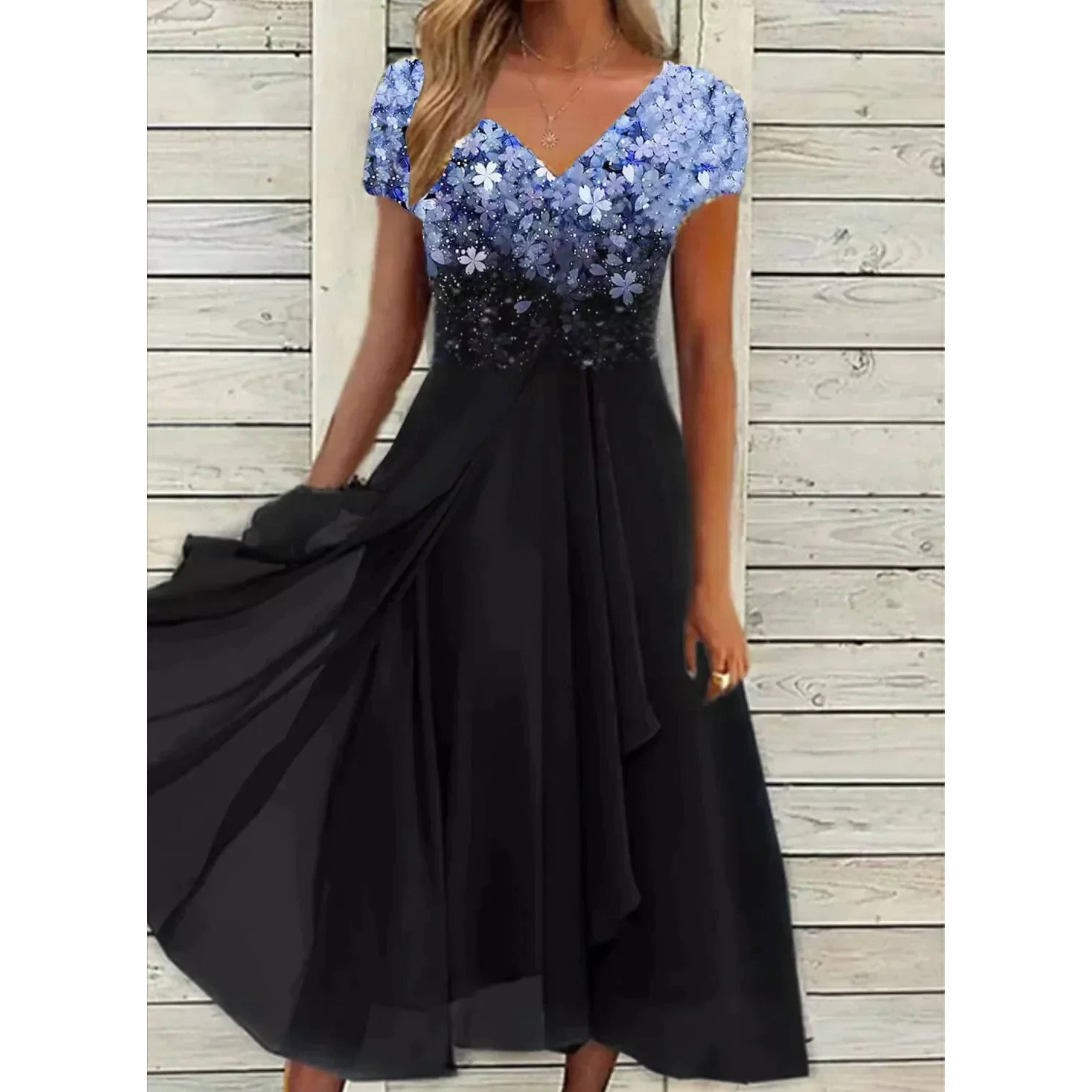 Robe femme élégante et polyvalente, longue et fluide, jupe noire avec haut floral bleu dégradé, col en V et manches courtes, portée devant un mur en bois blanc.
