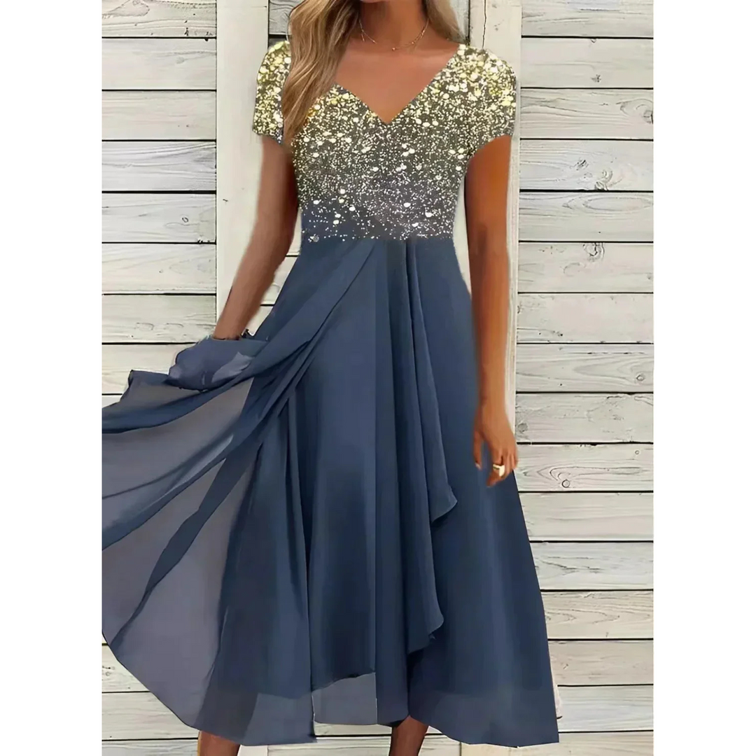 Robe femme élégante et polyvalente, longue et fluide, jupe bleu marine avec haut scintillant doré dégradé, col en V et manches courtes, portée devant un mur en bois blanc.